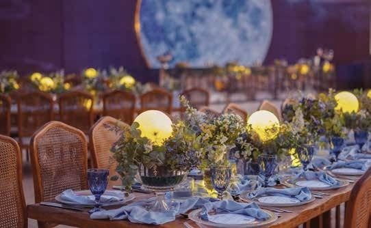 A long dinner table with blue linen, rattan chairs and glowing centrepieces beneath a moon backdrop.