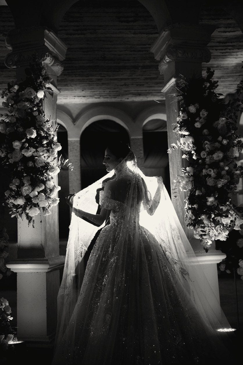 Black and white photograph of a bride in a full gown and veil beneath a floral archway.