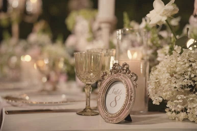 A candlelit table with gold-rimmed glassware and a framed table number among white flowers.